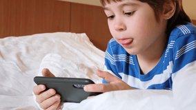 Portrait of a little boy playing online games on a smartphone, lying in bed at home. Child is using a mobile phone. Concept of gambling addiction. Gadget in hands. The tongue sticks out of the mouth.