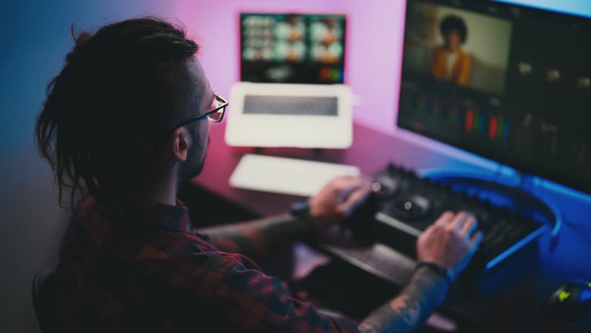 Man video editor wearing headphones sits at computer with backlight, looks in monitor. Video editing with music and color correction. Freelance, working in home office . Professional video processing. Royalty-Free Stock Footage #1100675483