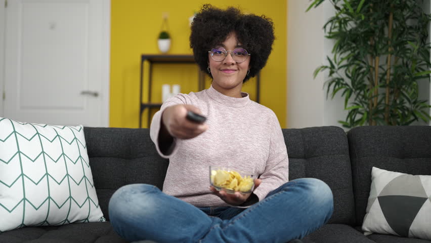 Young african american woman watching tv sitting on sofa eating chips at home