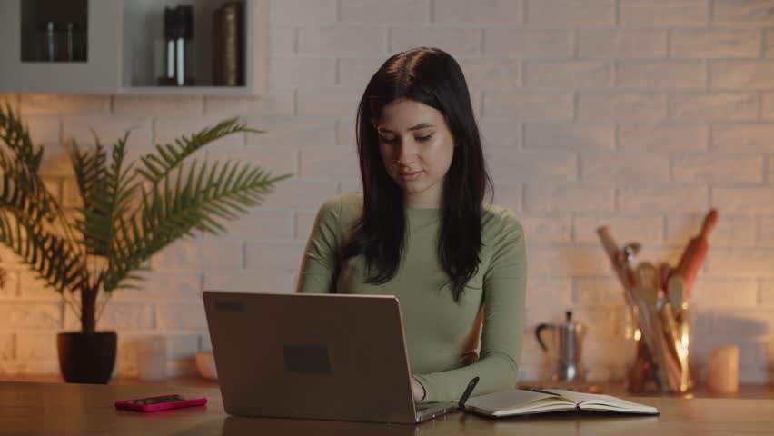 Freelancer woman working on a laptop at a desk in a home office. A student girl works at a computer and makes notes in a notebook | Shutterstock HD Video #1100683141