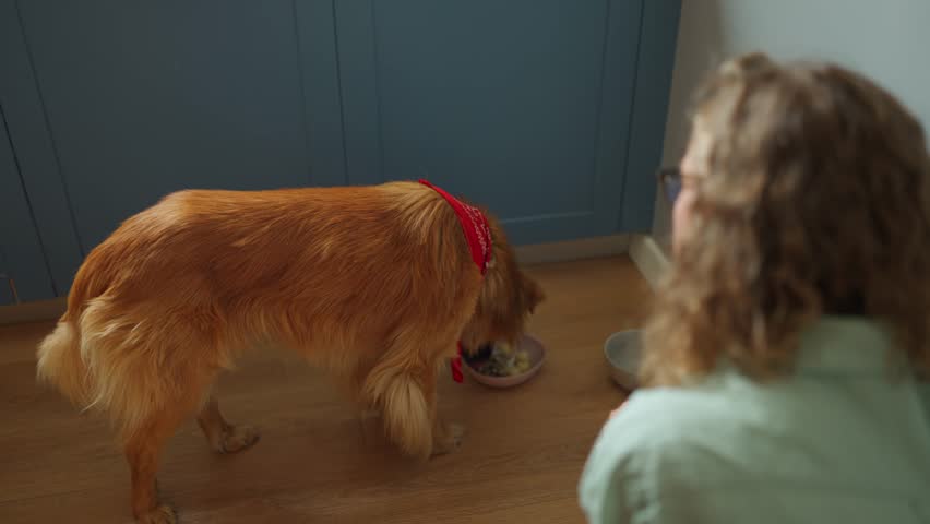 A curly haired woman watching a dog eat in the kitchen at home