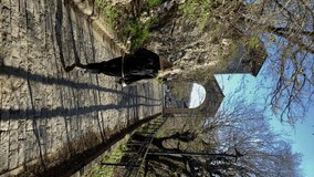 Vertical video. Blonde young woman in jacket along stone road to old arc of monastery. Motsameta, Georgia. Daylight, blue sky. 4K