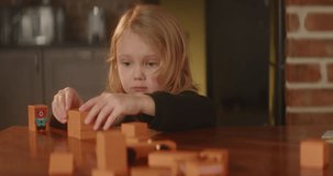 Bored sad girl plays busily on a table with orange wooden toy blocks, autistic child, cute kid builds street with houses at kitchen. Mental health problems. Nice home interior. High quality 4K footage