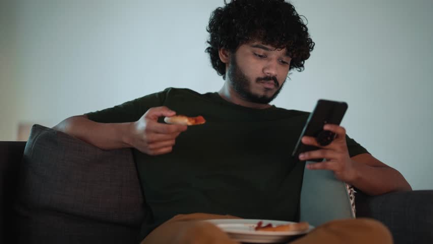 Pretty curly haired Indian man typing on mobile and eating pizza while sitting on the couch at home