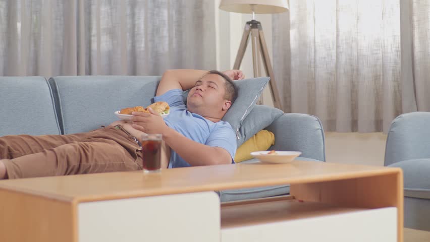 A Fat Asian Man Sleeping While Having French Fries With Ketchup Eating Fast Food On A Sofa In The Living Room At Home
