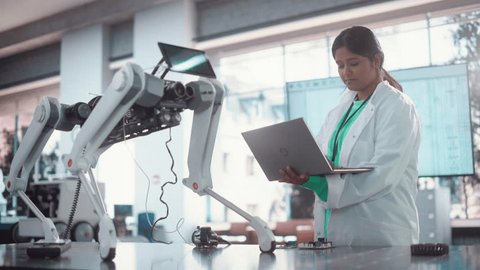 Young Indian Female Engineer Testing Industrial Programmable Robot Animal in a Factory Development Workshop. Professional Researcher in a Lab Coat Developing AI Canine Prototype, Using Laptop, videoclip de stoc