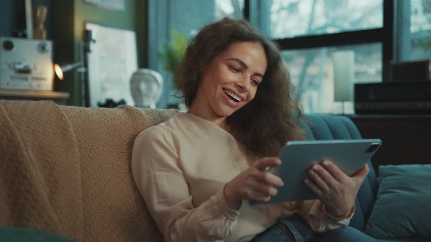 Footage of good-looking young relaxed woman sitting with digital tablet in hands, browsing through device. Cute girl watching funny video and chatting with friends in comfortable modern living room | Shutterstock HD Video #1100700397