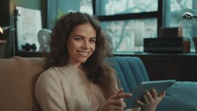 Footage of good-looking young relaxed Caucasian woman holding digital tablet in hands, connecting with others, posting on social media. Charming girl with long hair smiling at camera in cozy apartment - Powered by Shutterstock - Get 15% off with code: PIKWIZARD15