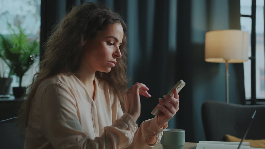 Young Caucasian woman sitting at table with laptop, holding smartphone in hands and posting on social media. Footage of focused girl in elegant blouse typing message in device. Indoors | Shutterstock HD Video #1100700417