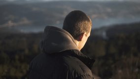 This stock slow motion video captures a young man's leisurely stroll in Domaio's Mirador do Monte do Faro on a sunny day. His black puffer jacket and hoodie make for a stylish and relaxed look.