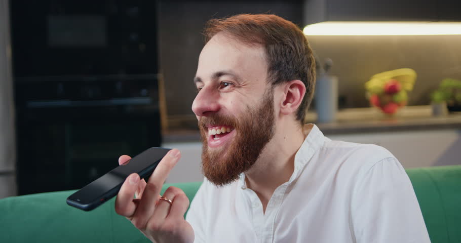 Successful freelancer man relaxing at home using mobile phone. Handsome young male recording voice audio message in social network while sitting on couch in kitchen. Communication, internet. | Shutterstock HD Video #1100705479