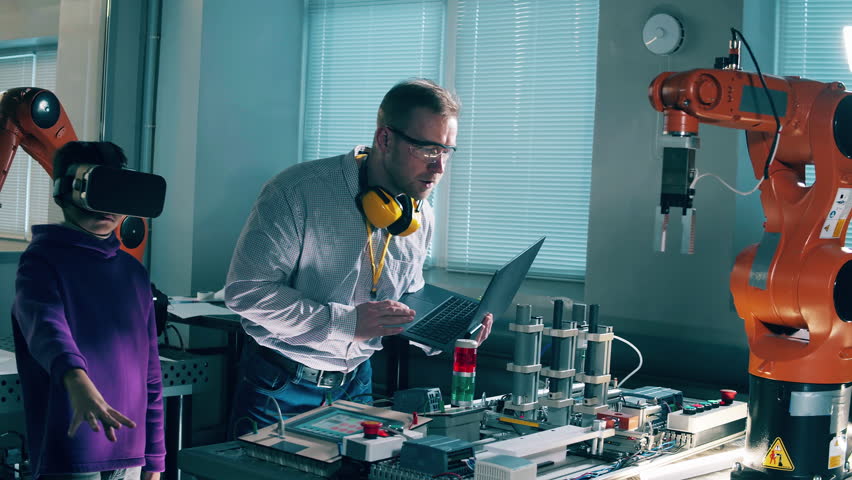 Engineer helps a boy in VR-glasses to operate a robotic arm