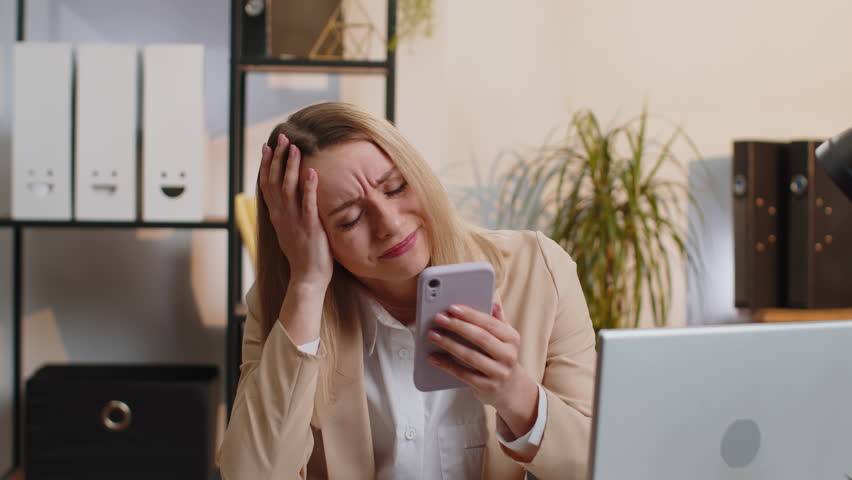 Young businesswoman in suit use smartphone typing browsing, loses becoming surprised sudden lottery results, bad news, fortune loss fail at home office desk workplace. Blonde freelancer worker woman | Shutterstock HD Video #1100707967