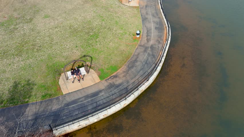 Overhead aerial footage of a couple in a swing and a family walking along a footpath along the rippling waters of the lake at Rhodes Jordan Park in Lawrenceville Georgia USA