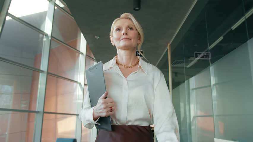 Slow motion mature confident woman 50s walking in corridor. Serious business woman going with folder in business center hallway. Portrait of focused businesswoman walking on meeting in office corridor