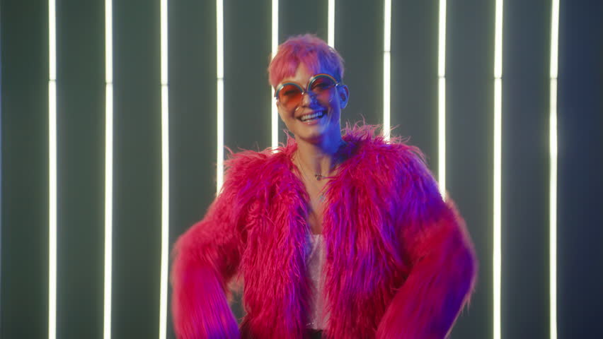 Funny smiling woman with pink hair having fun, smiling, dancing in studio against neon strip lights background. Funny girl in bright hot pink fur coat and sequin corset dancing by music in slow motion