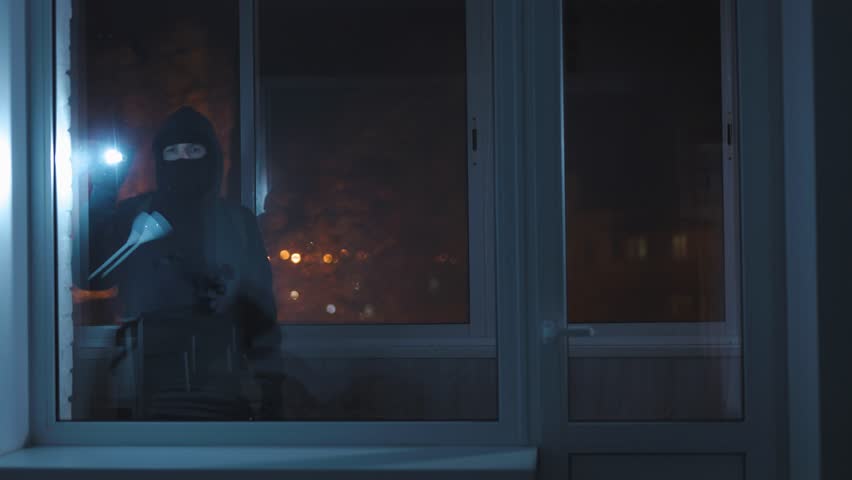 An unrecognizable burglar with a flashlight looks inside the house through the window. A male thief wants to break in and commit a theft during the night. Royalty-Free Stock Footage #1100727463