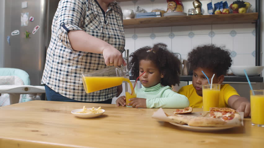 Babysitter feed two African-American kids with lunch. Preteen brother and sister eat pizza and juice for dinner. Caucasian foster mother give lunch to African-American son and daughter. Realtime.  | Shutterstock HD Video #1100728013