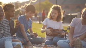 Diverse teenage children have picnic and play guitar in park. Multiethnic kids friends eat and play ukulele having fun outdoors. Group of kids on picnic. Childhood, friendship concept. Realtime.  - Powered by Shutterstock - Get 15% off with code: PIKWIZARD15