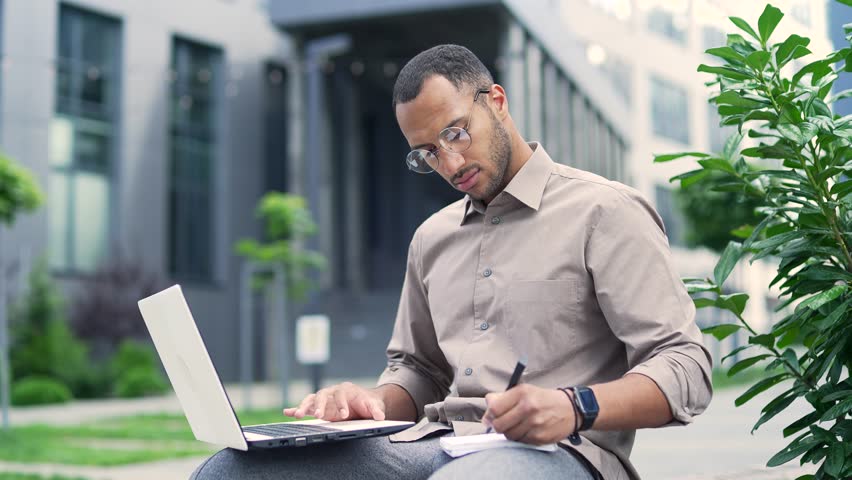 Young adult man works on a laptop while sitting on a bench on the street near an office building. Serious handsome mixed race male in a shirt and glasses writes in a notebook while browsing the device | Shutterstock HD Video #1100731841