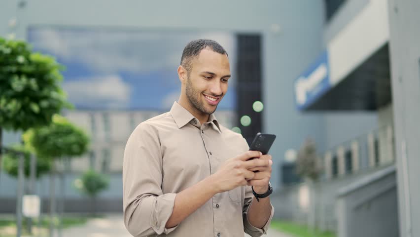 A smiling young adult man is using a smartphone while standing on the street near an office building. Happy handsome mixed race male checking email, chatting online, browsing social media, texting | Shutterstock HD Video #1100731849