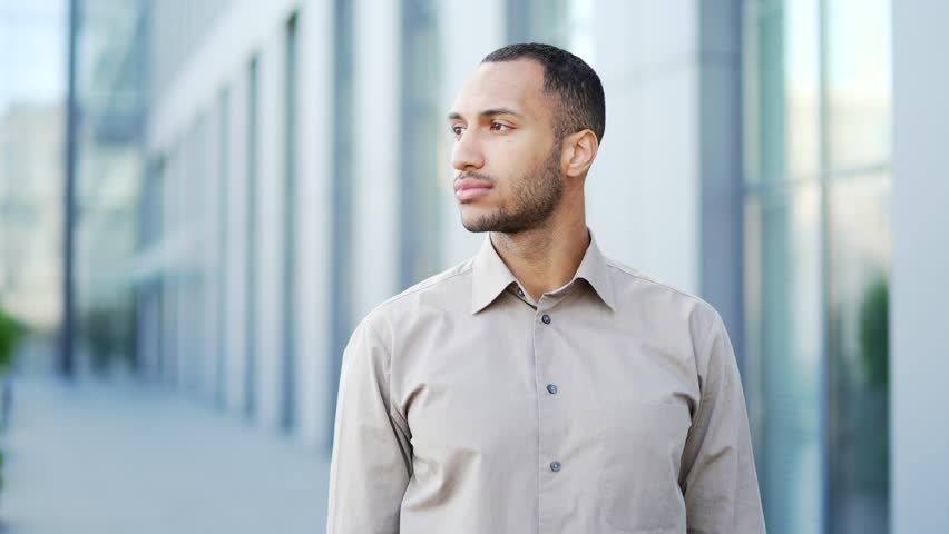 Portrait of a young adult man in a shirt smiling and looking at the camera while standing on the street near an office building. Happy and friendly handsome mixed race male posing. Head shot | Shutterstock HD Video #1100731889