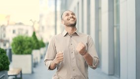 An excited adult young man celebrates victory, walking the street near the office building. A happy beautiful businessman jumps, dances, showing her hands a triumphant gesture. The concept of success - Powered by Shutterstock - Get 15% off with code: PIKWIZARD15