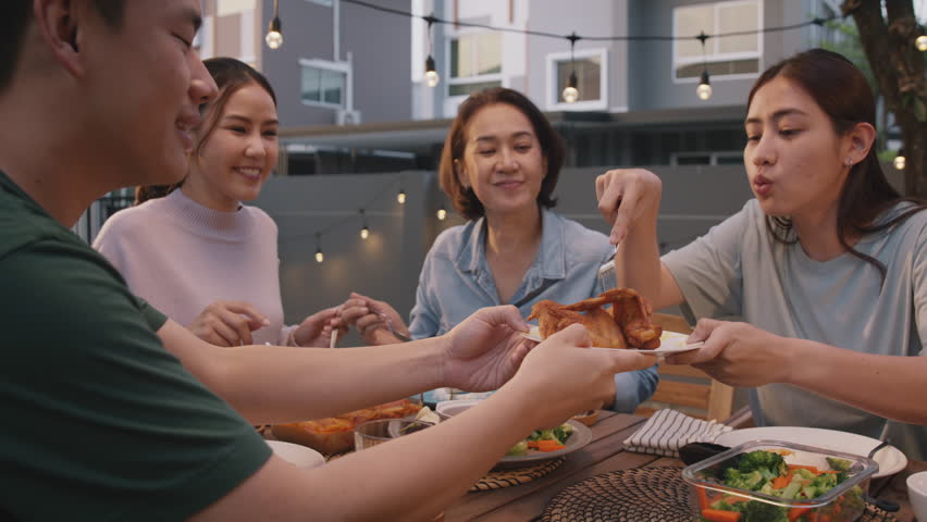 Mom enjoy thai meal cooking for family day home dining at dine table cozy patio. Mum passing serving food to group four asia people young adult man woman friend fun joy relax warm night picnic eating. | Shutterstock HD Video #1100732633