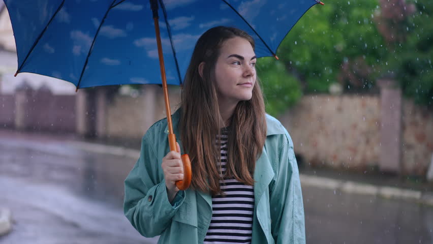 Joyful young woman standing in rain putting down umbrella smiling. Portrait of confident cheerful Caucasian lady posing and looking around on city street on rainy day