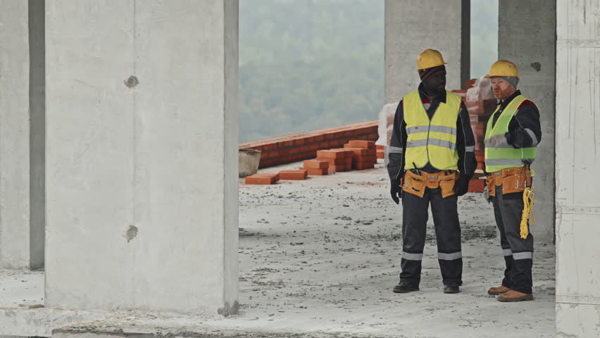Panning long shot of two ethnically diverse co-workers in workwear and safety helmets standing in building under construction discussing something
