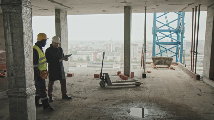 Mature Caucasian foreman and young adult Black builder walking along construction site discussing work plan | Shutterstock HD Video #1100732991