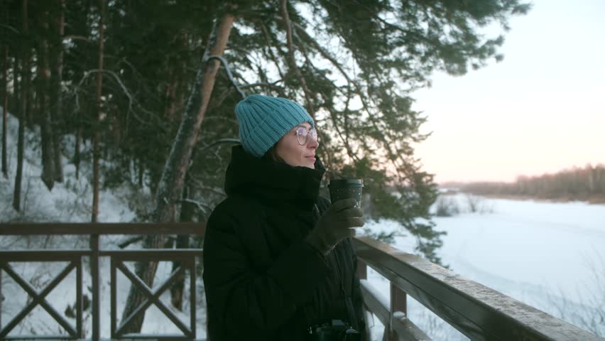 Happy smiling young woman standing on observation deck watching sunset holding in hand up with hot coffee. Tracking shot of charming lady drinking warming beverage outdoors in sunny winter day. High