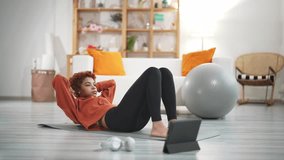 Woman lying on exercise mat at home and doing abs, watching fitness online video on digital tablet. At background sports equipment, fitness ball in living room. House workout and fitness without gym.