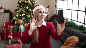 Young blonde woman having video call sitting on sofa by christmas tree at home
