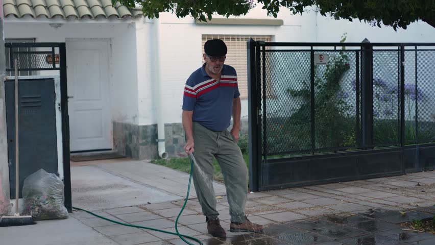 Retired mature man watering sidewalk in his house while whistles a song. South American retired people