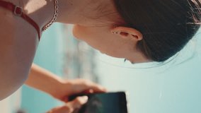 VERTICAL VIDEO: Close-up cute girl takes photos, videos of the seaport on a mobile phone. Girl uses social networks and streaming services on a smartphone when shooting yachts and ships