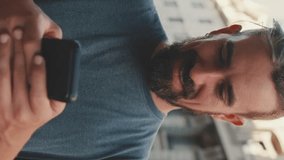 VERTICAL VIDEO, Young smiling man with beard stands on street using cellphone