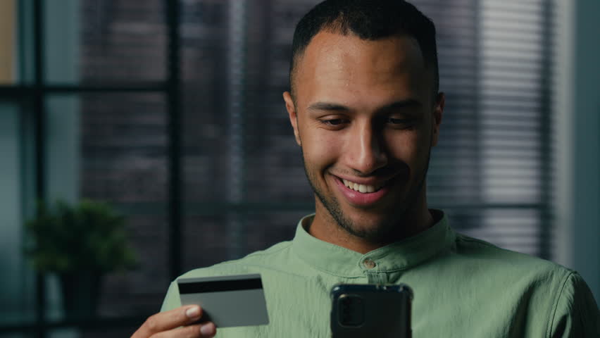 Smiling young african american man indoors holding credit card paying using mobile banking app on phone to buy online happy male customer shopper making purchase on smartphone pay with instant payment Royalty-Free Stock Footage #1100745805