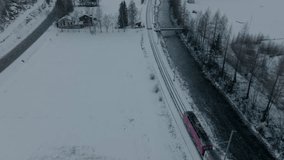 Aerial drone video of red train in Switzerland riding through stunning snowy winter landscape next to flowing river with alpine mountains panorama; clear blue sky; cute small Swiss mountain village