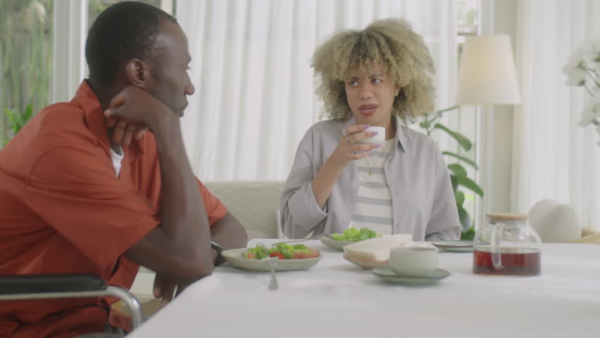 African American man with disability sitting in wheelchair, eating dinner and talking with wife while spending day together at home
