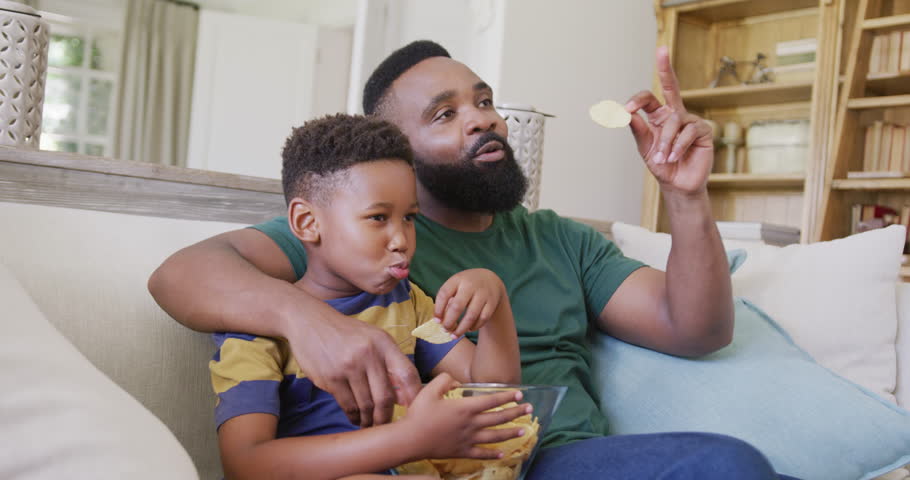 Happy african american father and son sitting on sofa, watching tv and eating crisps, in slow motion. Spending quality time, domestic life and childhood concept.