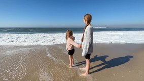 Two little sisters are playing with the waves on the beach of the Atlantic Ocean. Family time or vacation concept. 4K resolution video. High-quality 4k footage
