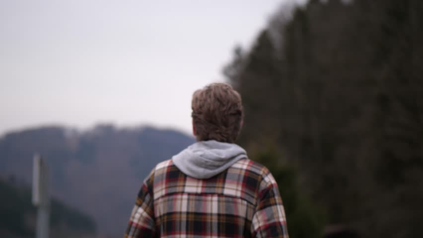 A man dressed in a plaid sweater is shown from behind taking a walk. In the background is a beautiful autumnal landscape with wooded hills to see