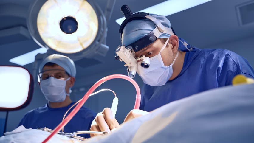 Intent look of a surgeon through the device glasses on the operated area. Doctor using the instrument carefully. Assistant at backdrop. Low angle view. | Shutterstock HD Video #1100773529