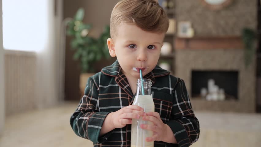 Portrait of cute smiling caucasian toddler boy who drinks milk through a straw. A child in pajamas enjoys drinking yogurt from a glass bottle through an ecological stainless steel drinking straw