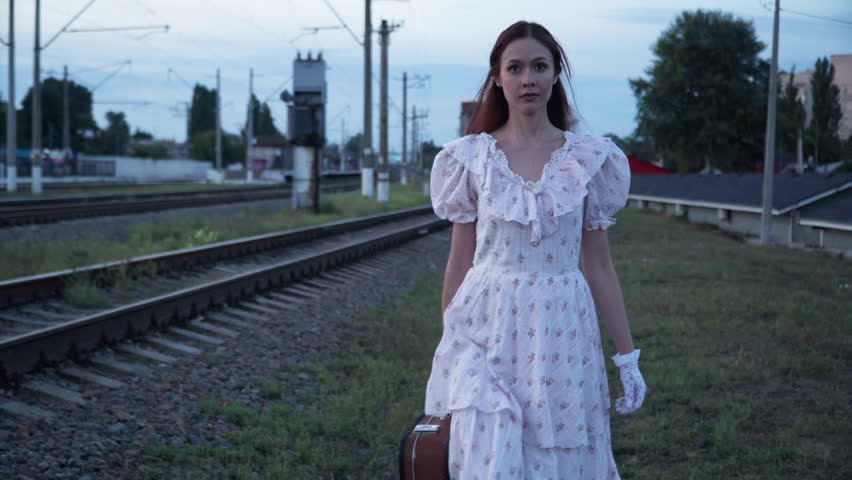 Dolly shot young confident woman in retro dress walking at railroad lines leaving. Front view portrait of beautiful slim Caucasian lady in vintage outfit with suitcase outdoors. Time travel cosplay Royalty-Free Stock Footage #1100781199