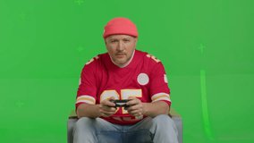 Close-up of a white man dressed in a red sports jersey and beanie sitting in a chair and playing with a joystick in a video game on TV on a green screen background, chroma key, front view