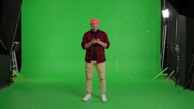 Full body video shot of caucasian man dressed in casual clothes and beanie, communicating on a mobile phone, texting messages, sending photo, reading text, standing on a green screen background