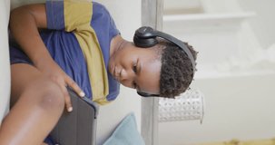 Vertical video of african american boy sitting on sofa and using tablet, in slow motion. Spending quality time, domestic life and childhood concept.