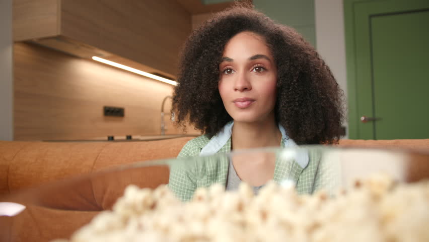 Woman Eats Popcorn Close Up. Smiling mixed race woman enjoys TV watching sitting on couch with popcorn bowl. Royalty-Free Stock Footage #1100786029
